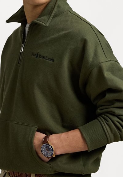 Olivgrön zip-up sweatshirt med hög krage och broderad logotyp. Har sidofickor och texturerade stickade ärmslut. Bärs med en armbandsur.