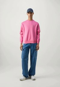 En ung man står klädd i en rosa sweatshirt, blå cargobyxor, vita sneakers och en mörk keps mot en enfärgad ljus bakgrund.