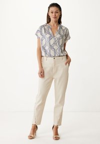 Camicia a maniche corte con un motivo geometrico blu e bianco, abbinata a pantaloni beige chiaro a gamba affusolata con dettagli laterali in laccatura. Sandali con tacco alto dorati.