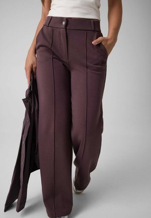 Person trägt hoch taillierte, weit geschnittene dunkelbraune Hose und weißes Oberteil, hält eine passende dunkelbraune Jacke in einer Hand.