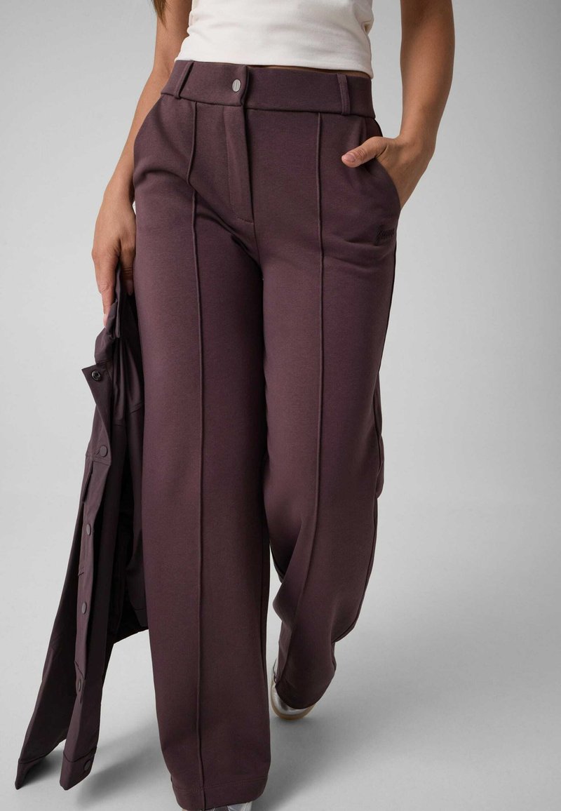 Personne portant un pantalon large taille haute marron foncé et un haut blanc, tenant une veste marron foncé assortie dans une main.