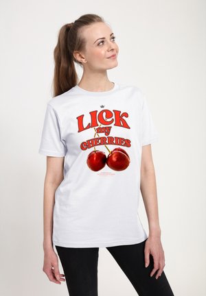 Kvinde med brunt hestehale iført hvid T-shirt med store kirsebær og rød tekst "Lick my cherries," stående foran en ensfarvet lys baggrund.