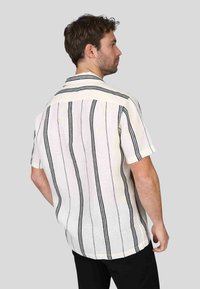 Chemise à manches courtes, col classique en tissu blanc texturé avec des rayures verticales noires, présentant une coupe décontractée et un ourlet arrondi.