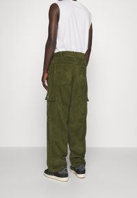 En person som bär olivgrön kordflodddokcargo byxor och svarta Adidas sneakers, står mot en enkel vit bakgrund, sedd bakifrån.