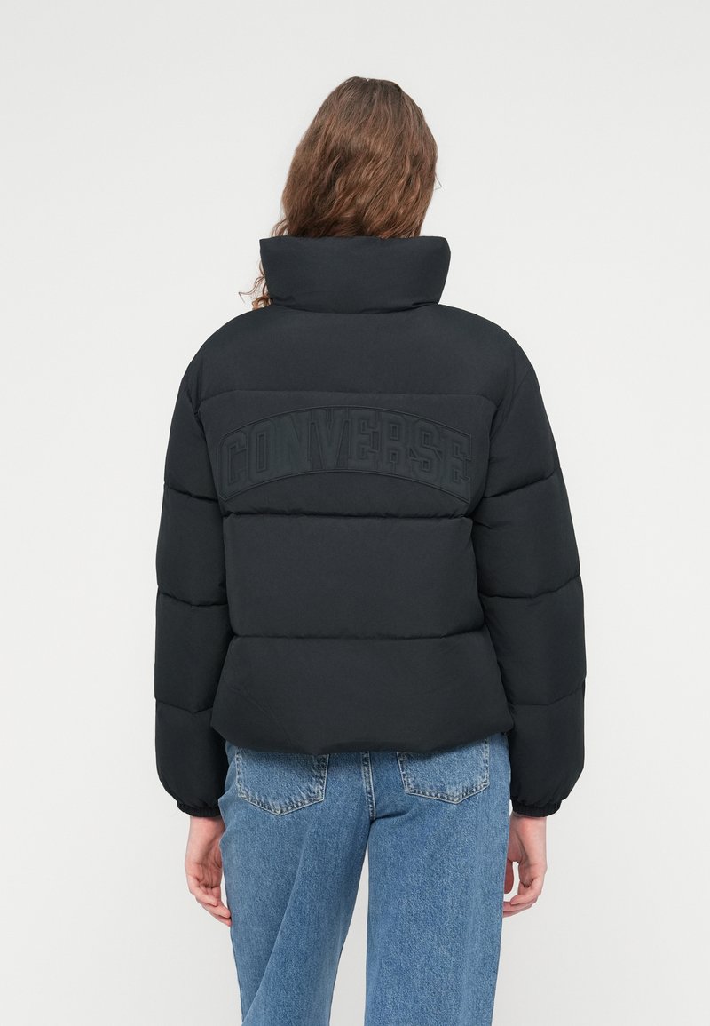 Sort dunjakke med høj krave, der har et stort præget "CONVERSE"-logo på ryggen. Jakken har en firkantet pasform og elastiske manchetter.
