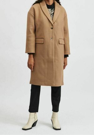 Personne portant un manteau camel mi-long, un pantalon noir, des bottines blanches et un pull à motifs en dessous.