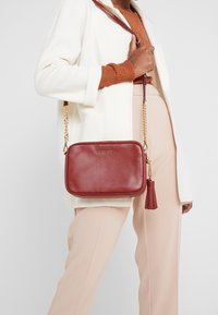 Kobieta w białym płaszczu i beżowych spodniach trzyma bordową skórzaną torebkę na ramię z złotym łańcuchem i detalem w postaci frędzla.