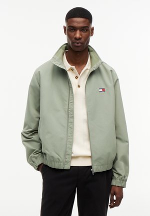 Muž v svetlo zelenej bunde na zips cez krémovú košeľu na gombíky a čierne nohavice, stojaci s rukami v vreckách na bielom pozadí.