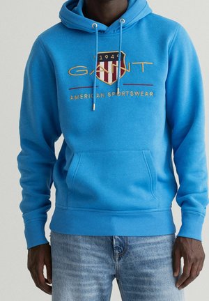 Homme portant un sweat à capuche GANT bleu vif avec logo et texte « American Sportswear » associé à un jean bleu sur fond uni.