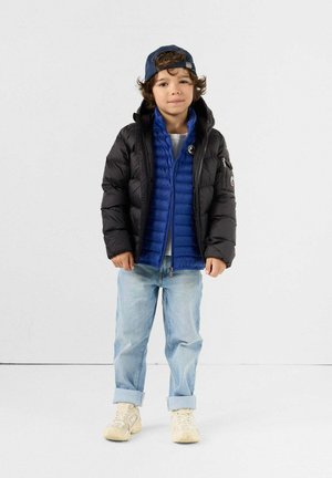 Young boy wearing a black puffer jacket over a blue jacket, light blue rolled-up jeans, white sneakers, and a backward blue cap standing.