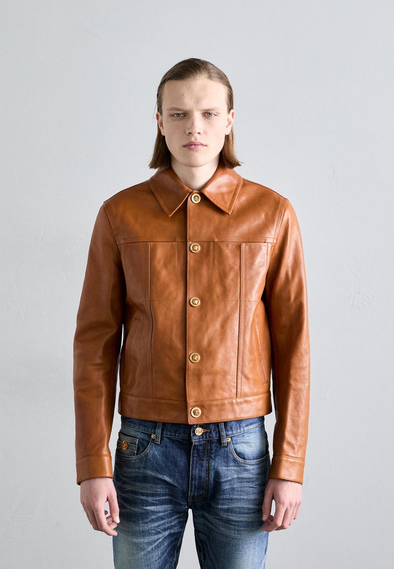 Braune Lederjacke mit kragenförmigem Hals, ausgestattet mit goldenen Knöpfen und zwei Fronttaschen. Trägt man über blauen Jeans mit verwitterter Textur.