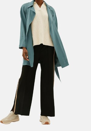 Personne portant un trench-coat bleu sarcelle, un pull en maille crème à col en V, un pantalon noir à jambes larges avec des bandes latérales, et des baskets beiges sur fond blanc.