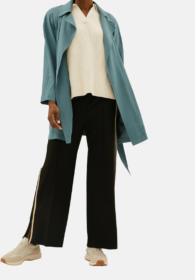Personne portant un trench-coat bleu sarcelle, un pull en maille crème à col en V, un pantalon noir à jambes larges avec des bandes latérales, et des baskets beiges sur fond blanc.