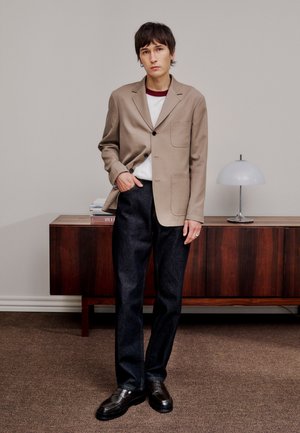 Tânăr purtând un sacou bej, cămașă albă, blugi închiși la culoare și pantofi negri, stând în fața unei comode din lemn cu o lampă albă.