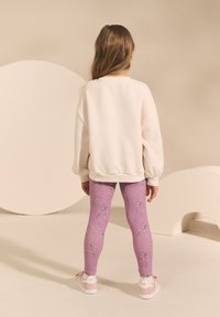 Svijetlosmeđa majica dugih rukava, ljubičaste helanke s uzorkom kontrastnih mašni i ružičaste tenisice s bijelim detaljima; glatki materijal i opušteni kroj.