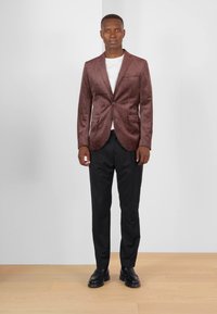 Brauner Samtblazer mit einem einzigen Knopf, mit Revers, zwei Taschen, kombiniert mit schwarzen Hosen und schwarzen Schuhen vor einem neutralen Hintergrund.