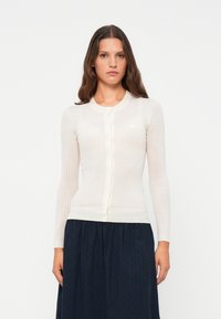 Light ribbed cardigan in off-white, featuring a round neckline, long sleeves, and front buttons. Worn with a dark pleated skirt.
