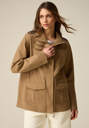 Kobieta ubrana w beżową kurtkę użytkową z klapkowymi kieszeniami i regulowanymi ściągaczami, trzymająca częściowo zapięty zamek przy kołnierzu.