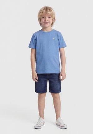 Ξανθός αγόρι που στέκεται, φορά μπλε μπλούζα, σκούρο τζιν σορτς και λευκά αθλητικά παπούτσια, χαμογελώντας μπροστά σε απλό φωτεινό φόντο.