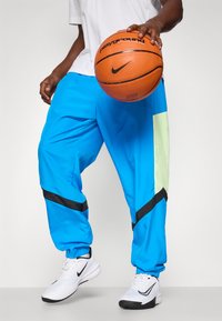 Blue athletic pants with black accents and a mesh green panel, paired with white shoes. The person holds a basketball.