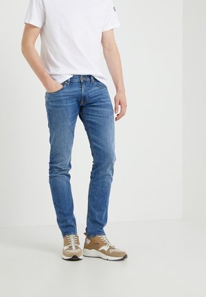 Mann steht mit einer Hand in der Tasche, trägt ein schlichtes weißes T-Shirt, blaue Jeans und beigefarbene Sneaker in einer minimalistischen Inneneinrichtung.
