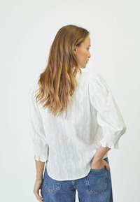Blusa bianca con motivi geometrici testurizzati, maniche a sbuffo e polsini elastici, abbinata a jeans blu. Vista posteriore mostrata.