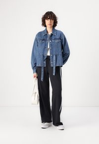 Veste en denim avec une coupe décontractée, couleur bleu clair, grandes poches avant et fermeture à boutons. Associée à un pantalon de jogging noir et un sac blanc.