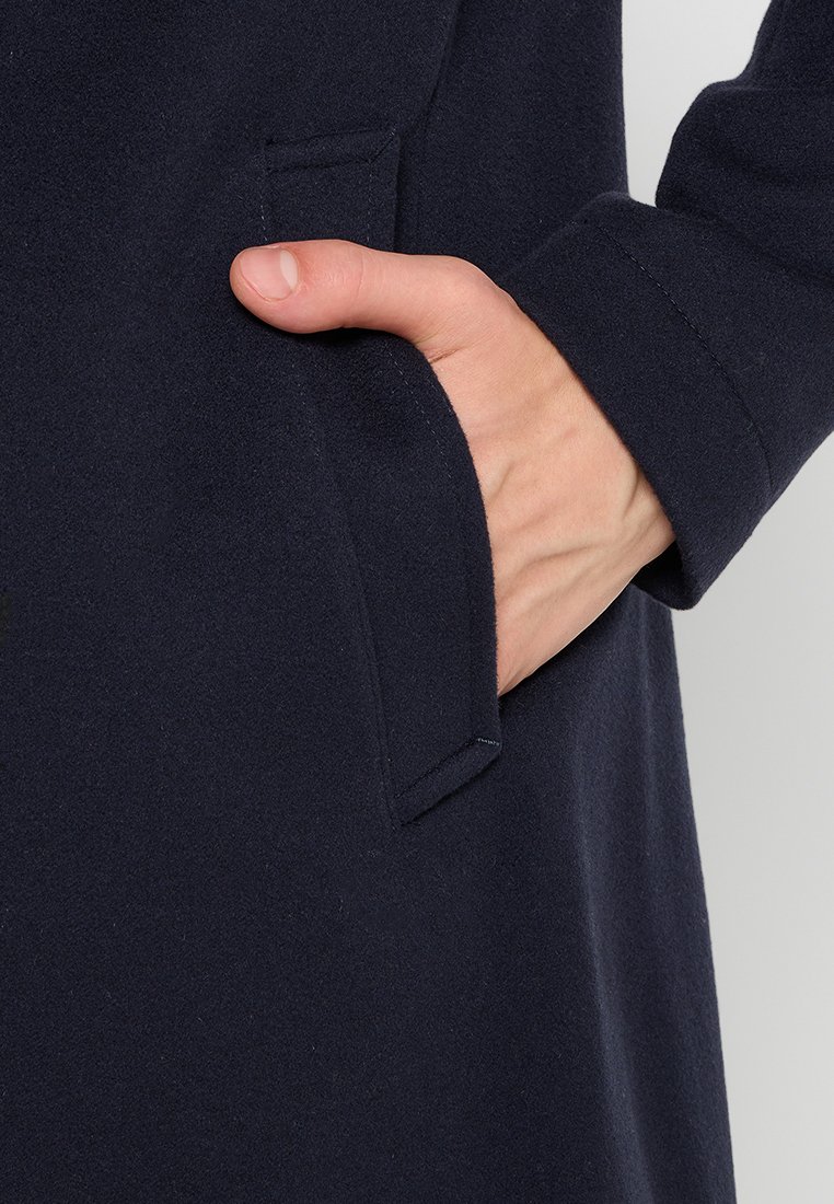 Main dans la poche latérale d'un manteau en laine bleu marine, montrant le poignet de la manche et la texture du manteau dans un éclairage neutre.