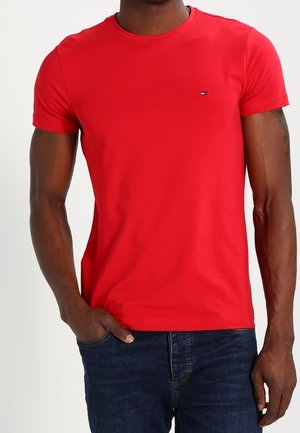 Homme portant un t-shirt rouge uni à manches courtes et col rond avec un petit logo sur la poitrine et un jean bleu foncé.