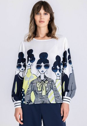 Vrouw draagt een witte blouse met een grafische print van gestileerde vrouwen met zonnebrillen en patronen, staande tegen een effen achtergrond.