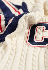 Kabelgebreide trui in crème met navy en rode gestreepte details. Heeft een geborduurd "C"-logo in navy omlijnd met rood. Zachte textuur.