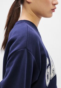 Tengerészkék pulóver kerek nyakkal és bordázott mandzsettákkal. Az elején nagy, világos színű "GAP" felirattal. Sima textúrájú.