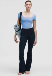 Blaues enges Oberteil mit kurzen Ärmeln und einem schwarzen Bund, kombiniert mit schwarzen, ausgestellten Yoga-Hosen. Trägt eine gerollte hellblaue Yogamatte.