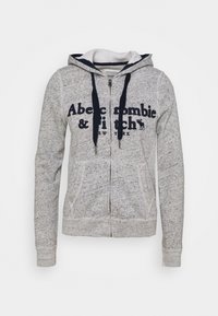 Heathergrå lynlåshættejakke med marineblå accenter, kængurulommer, dobbeltsyet hætte og frontlogo-tekst. Blødt, elastisk stof, ribstrikkede ærmeafslutninger.