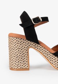 Sandalia de tacón de ante negro con plataforma trenzada beige, correa al tobillo con hebilla dorada y plantilla suave color canela. Diseño de tacón bloque robusto.