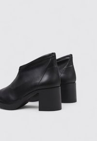 Une paire de bottines noires en cuir avec des talons carrés, des coutures minimales et une finition lisse sur fond gris clair.