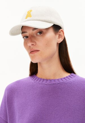 Jonge vrouw met sproeten, die een beige pet met geel logo en een paarse gebreide trui draagt, kijkt iets opzij, op een witte achtergrond.