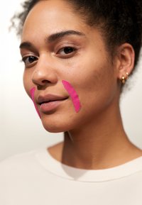 Gros plan d'un visage avec des bandes de tissu rose appliquées sur les joues, sur fond clair, avec une texture de peau naturelle et un éclairage doux.