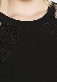 Mustapaita, jossa on pitsikoristeet olkapäillä ja teksturoituja paneeleja. Sileä kangas, pyöreä pääntie ja lyhyet hihat.