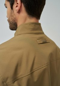 Veste tan avec un col haut et des détails de couture minimaux. Présente une étiquette carrée près du col, mettant en valeur une texture lisse et épurée.