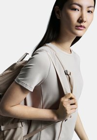 Camiseta deportiva beige de manga corta, llevada por una persona que sostiene una mochila ligera con correas de malla y detalles en acentos. Textura sedosa.
