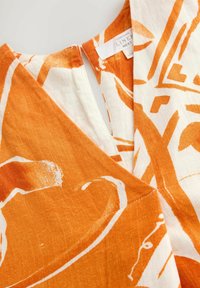 Hørblandet stof i orange og hvid med abstrakte mønstre. Har en V-udskæring og syningsdetaljer. Blød, tekstureret overflade.