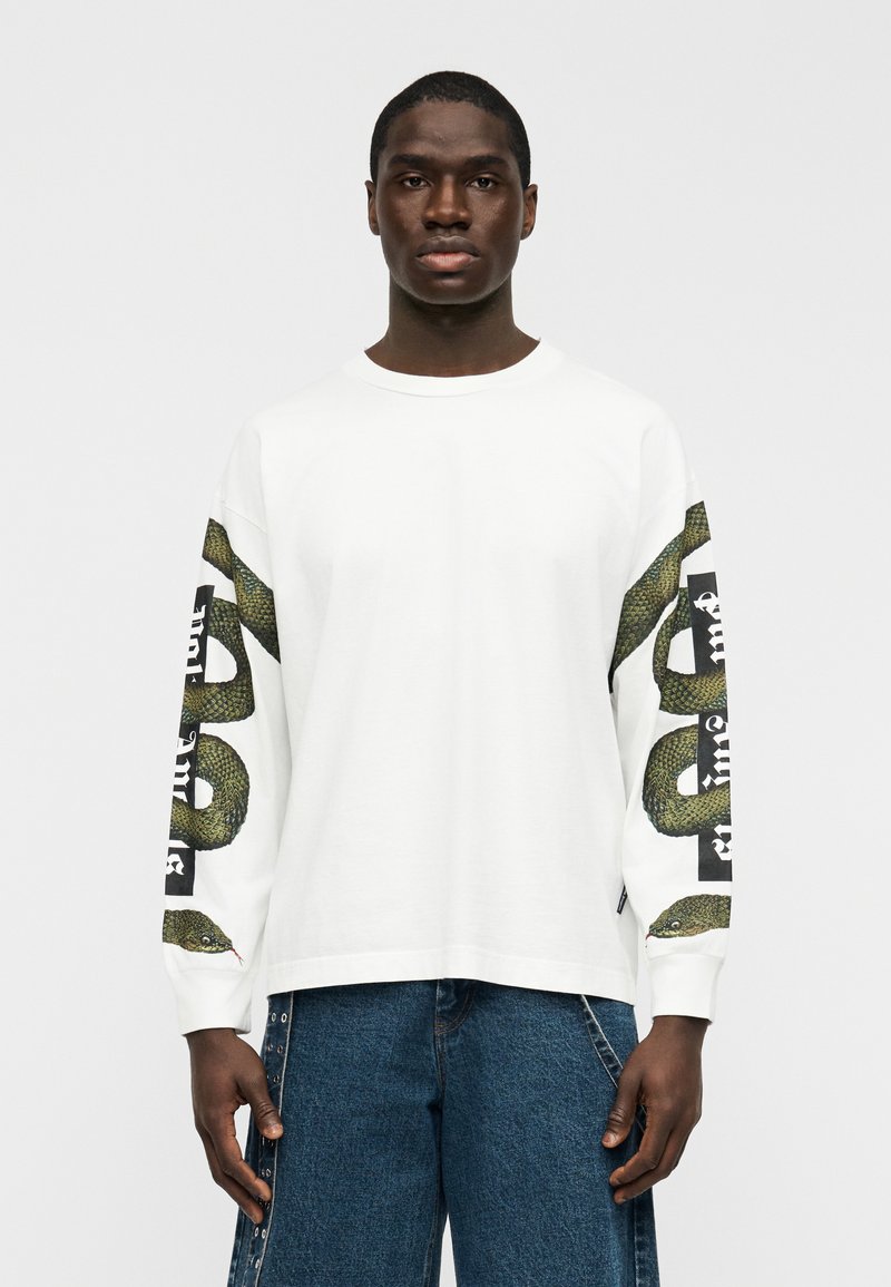 Λευκό βαμβακερό μπλουζάκι με μακριά μανίκια, που διαθέτει γραφική εικόνα πράσινου φιδιού και μαύρο κείμενο στα μανίκια· άνετη εφαρμογή με στρογγυλή λαιμόκοψη.