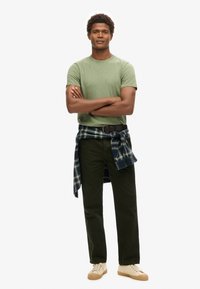 T-shirt verde a maniche corte, pantaloni verde scuro e scarpe chiare. Una camicia a quadri scura è legata in vita.
