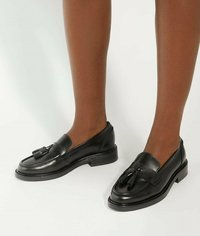 Black leather loafers with a smooth texture, tassel accents, rounded toe, and a low block heel, shown on bare legs against a neutral background.
