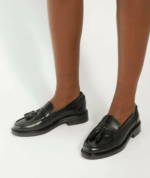 Zwarte leren instappers met een gladde textuur, franje-accenten, ronde neus en een lage blokhak, getoond op blote benen tegen een neutrale achtergrond.