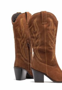 Botas de cowboy de ante marrón con punta afilada, tacón negro apilado y detalle de bordado de llamas amarillas en el caño. Cierre con cremallera en el lateral.