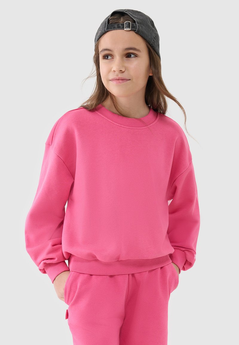 Jeune fille portant un sweat-shirt rose vif et un pantalon, les mains dans les poches, regardant légèrement à droite, portant une casquette sombre à l'envers.