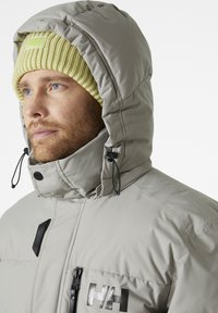 Lichtgrijze pufferjas met een capuchon, voorzien van zwarte accenten en een ritszak. Draag het met een geribde, lichtgele beanie.
