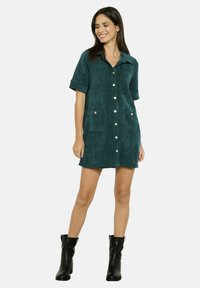 Abito camicia in corduroy verde acqua con maniche corte, chiusura frontale con bottoni, due tasche frontali e colletto, abbinato a stivali neri alla caviglia.